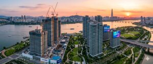 High-quality, photorealistic, 8k, cinema lighting, professional photography style. A panoramic aerial view of high-rise luxury apartment complexes under construction near the Han River in Gangdong-gu, Seoul. The scene features a blend of modern glass architecture and lush green parks, with the warm glow of sunset reflecting off the river and building facades, symbolizing urban transformation and future value.